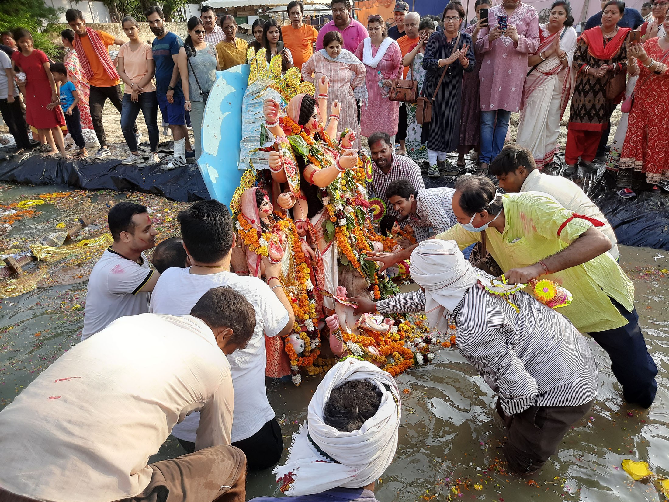 People stand on the edges of an artificial pond, watching a group of men as they prepare to lower a colourful idol of the Goddess Durga, laden with flowers, into the pond water